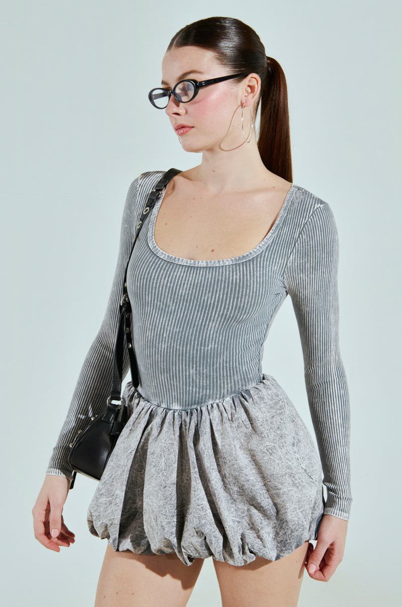 A person with a ponytail wears glasses and large hoop earrings. They are dressed in a GREY GIVE ME A LITTLE MINI DRESS. A black shoulder bag hangs on one arm against a light gray background.