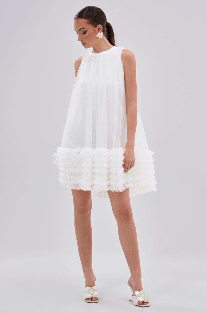 A woman wearing the ARI TULLE RUFFLE MINI DRESS IN WHITE stands against a plain light gray background, styled with white heeled sandals featuring floral details and statement earrings.