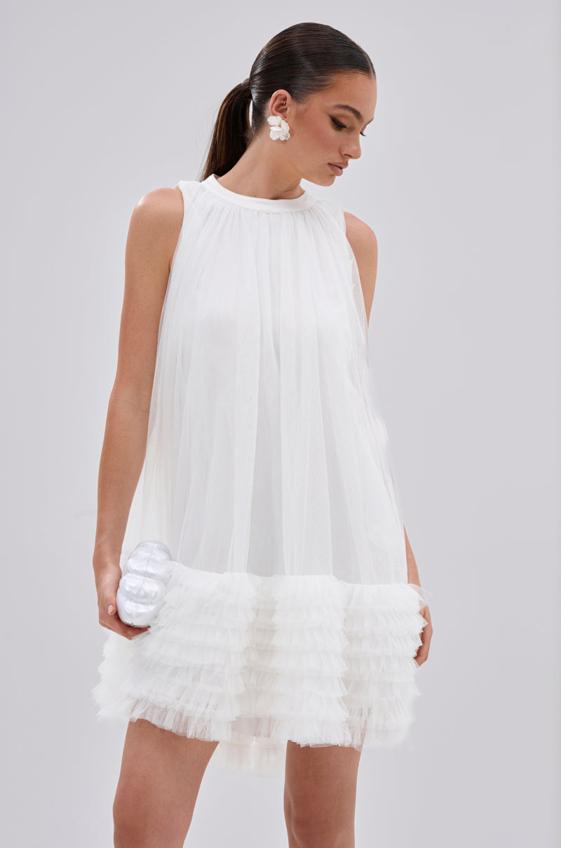 Wearing the ARI TULLE RUFFLE MINI DRESS IN WHITE, a woman accessorizes with large white earrings and a small white clutch. Her hair is styled in a sleek ponytail.