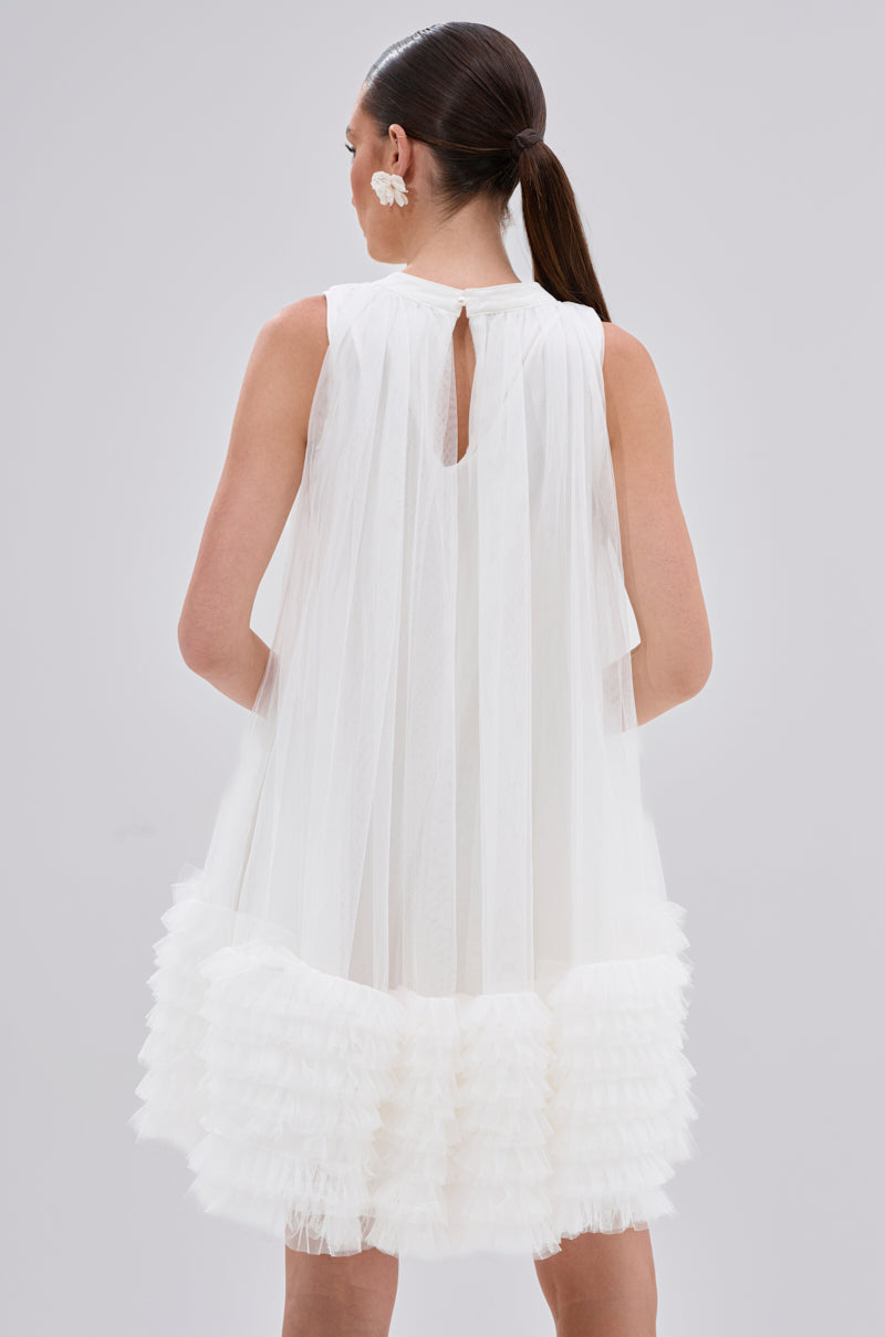 A woman with long brown hair in a low ponytail is shown from the back wearing the ARI TULLE RUFFLE MINI DRESS IN WHITE, featuring sheer accordion-pleated tulle and ruffled layers at the hem. She pairs it with a white floral earring against a plain background.