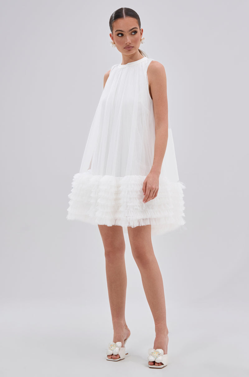 A woman wears the ARI TULLE RUFFLE MINI DRESS IN WHITE, a sleeveless mini dress with a flowy silhouette and ruffled hem, paired with white open-toe heels with floral accents. Her hair is pulled back as she poses against a plain background.