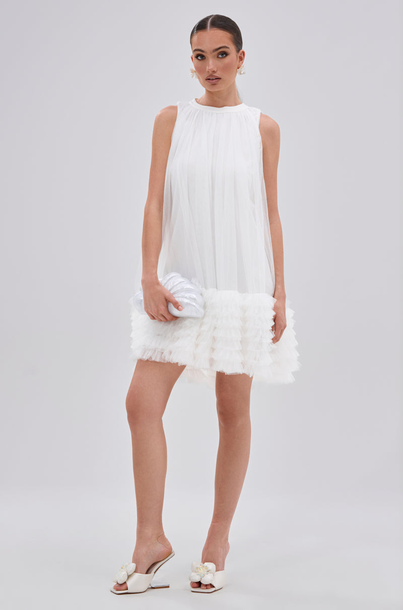 A woman in the ARI TULLE RUFFLE MINI DRESS IN WHITE, featuring a sheer overlay and ruffled hem, holds a silver clutch. She pairs it with white heeled sandals with bows, her hair styled back, against a plain light background.
