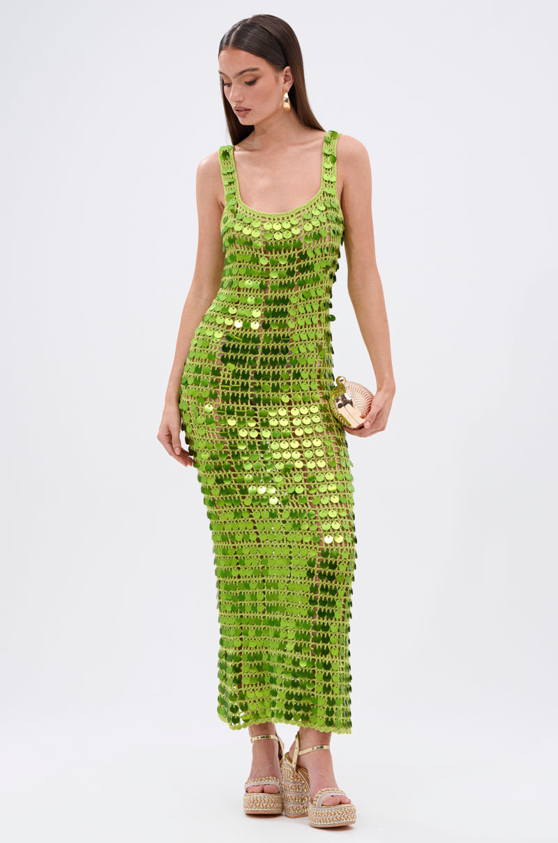 A woman in the SHINE BRIGHT CROCHET SEQUIN MAXI DRESS IN GREEN, form-fitting and sleeveless, with a clutch and platform sandals, poses against a white background—ready to light up any occasion.