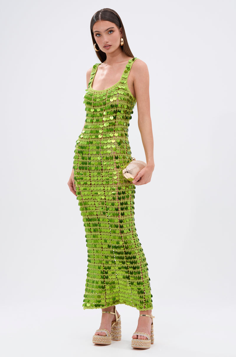 A woman in the SHINE BRIGHT CROCHET SEQUIN MAXI DRESS IN GREEN, styled with a woven clutch and platform sandals, stands against a plain white background—poised to dazzle.