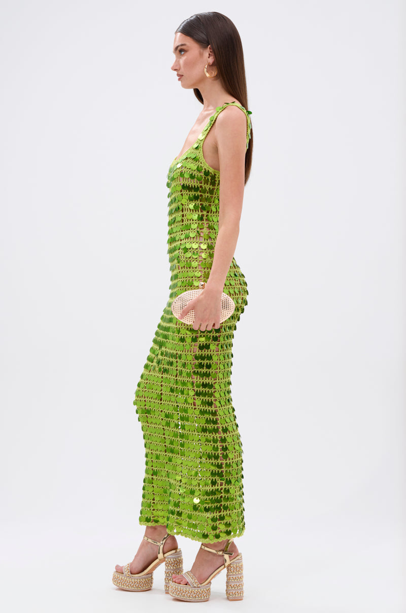 A woman in profile wears the SHINE BRIGHT CROCHET SEQUIN MAXI DRESS IN GREEN with platform woven sandals and a textured clutch, shining against a plain white background.