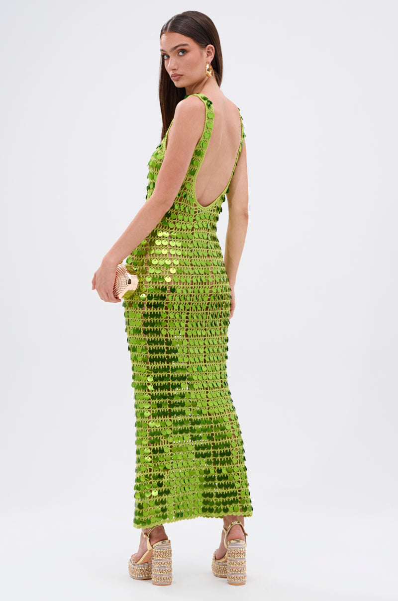 A woman stands sideways, wearing the SHINE BRIGHT CROCHET SEQUIN MAXI DRESS IN GREEN with large sequins and a low back. She holds a small clutch and wears platform sandals, standing out against the plain white background.