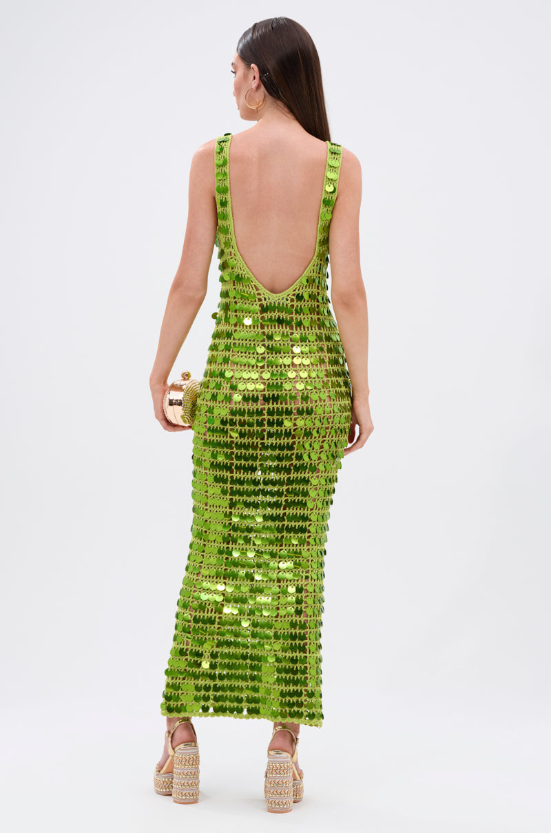 A woman stands facing away in the SHINE BRIGHT CROCHET SEQUIN MAXI DRESS IN GREEN with a low scoop back, holding a gold clutch and wearing platform sandals. Her long straight hair cascades down her back.