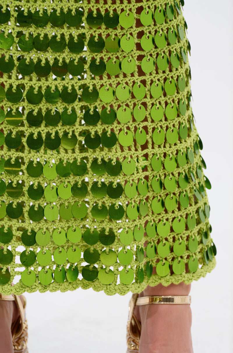 Close-up of the SHINE BRIGHT CROCHET SEQUIN MAXI DRESS IN GREEN, featuring overlapping shiny green sequins with scalloped edges, shown with gold high-heeled sandals and part of a person’s lower legs.