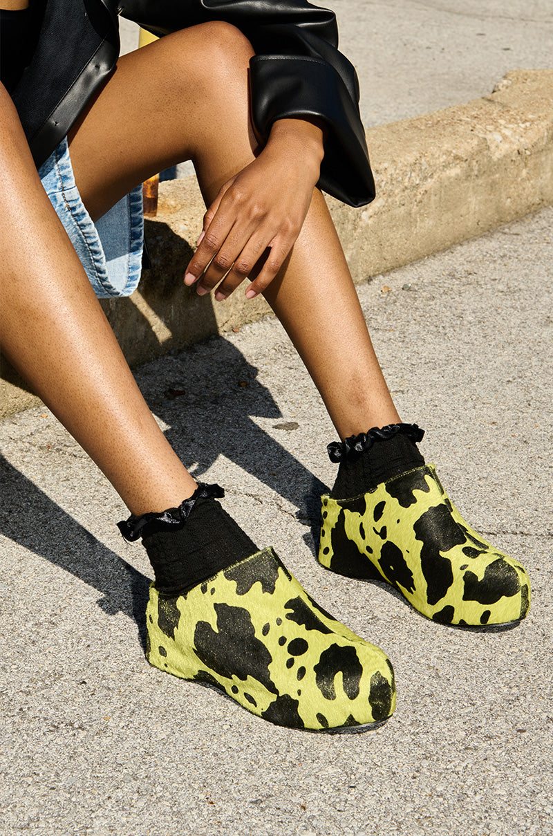A person is sitting on a concrete block, wearing a black jacket, light blue denim shorts, and bright yellow clogs with black cow print patterns. The individual also sports black socks with frilled tops. Their AZALEA WANG DARWIN LIME FLATFORM clogs feature a synthetic pony hair upper against the concrete pavement and curb backdrop.