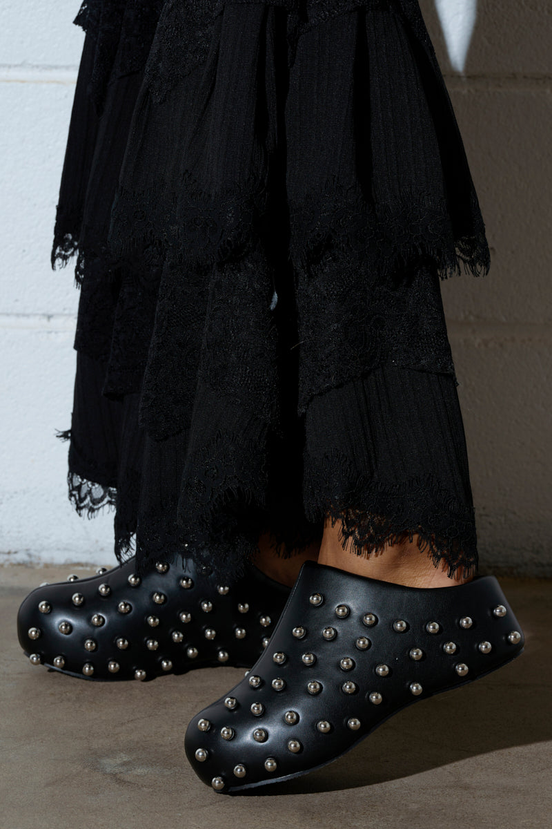 Against a white brick wall, someone stands on a concrete floor wearing a long black skirt with lace details and AZALEA WANG DARWIN STUD FLAT CLOGS IN BLACK, featuring vegan leather and metal studs with a flatform sole.