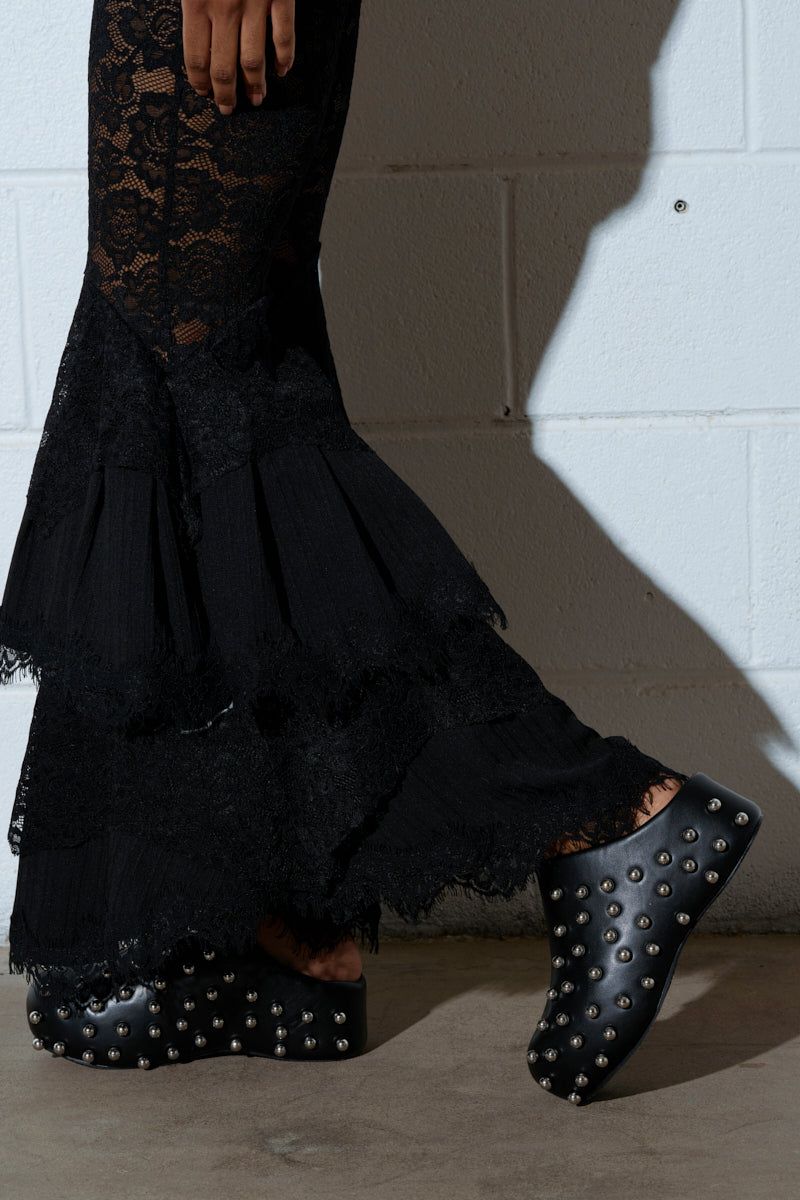 A person wearing black lace flared pants and AZALEA WANG DARWIN STUD FLAT CLOGS, crafted with vegan leather and embellished with silver studs, stands against a white textured wall.