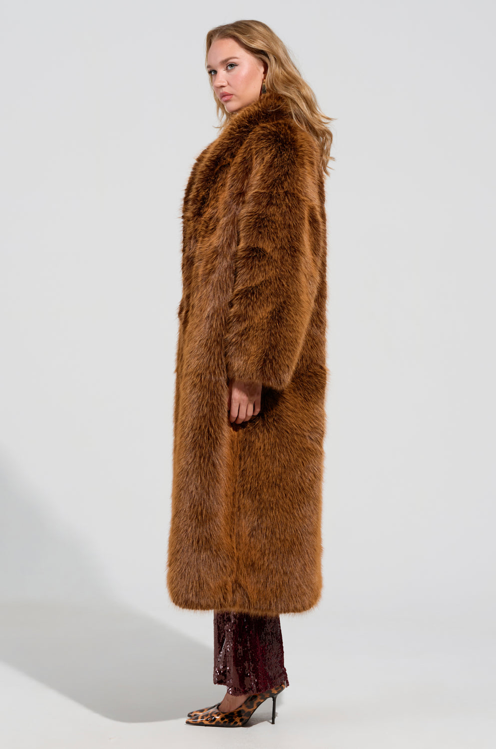 A person wearing the DAYANNE FAUX FUR TRENCH IN BROWN, featuring a rich brown hue and an oversized silhouette, stands in profile, looking forward as their long wavy hair cascades down. Patterned heels and dark patterned pants complete the ensemble against a plain background.
