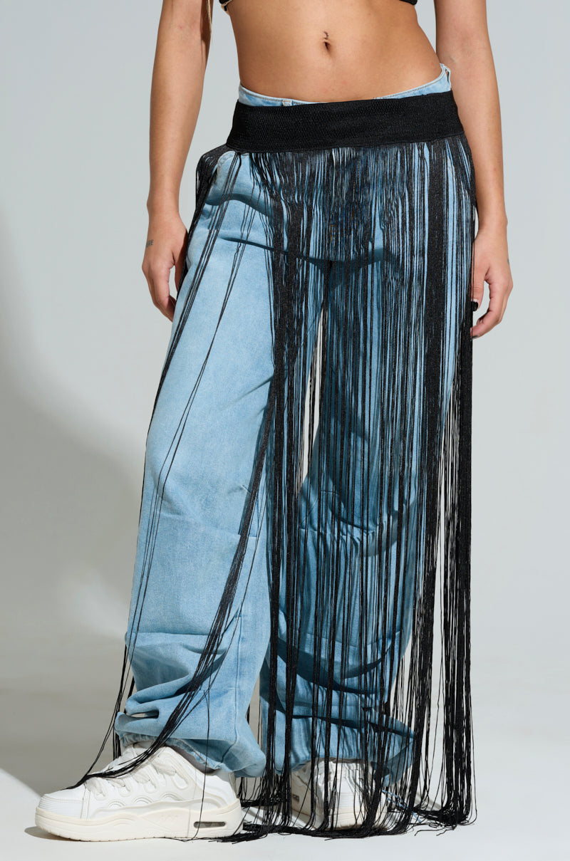 A person wearing light blue jeans and white sneakers is styled with the SO FREE FRINGE BELT SKIRT, featuring a black, cascading fringe detailing that drapes to the floor, partially revealing the jeans underneath. The background is a simple, light grey.