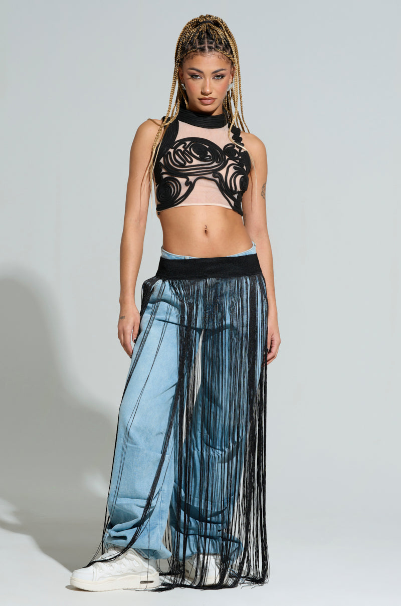 The individual with braided hair sports a chic sheer top adorned with intricate patterns, paired with loose denim pants and a black skirt overlay featuring cascading fringe. They exude confidence against a simple backdrop, showcasing a modern and edgy fashion sense highlighted by the SO FREE FRINGE BELT SKIRT.