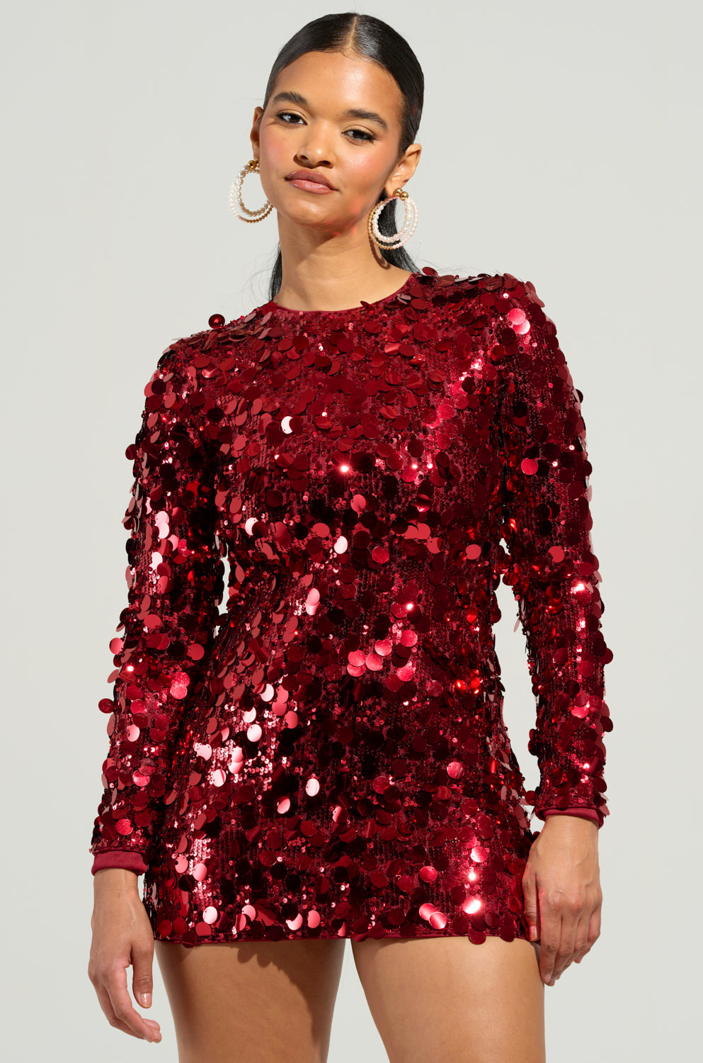 A person stands confidently in the SHE'S ALL THAT SEQUIN MINI DRESS with long sleeves, exuding a party-ready vibe as they pair it with hoop earrings and neatly styled hair against a plain background.