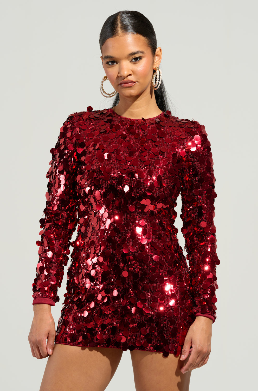 A person stands against a plain background in the SHE'S ALL THAT SEQUIN MINI DRESS, a radiant, shiny red sequin mini dress featuring a bodycon fit and long sleeves. With straight black hair and hoop earrings, they exude a party-ready ensemble vibe while looking directly at the camera.