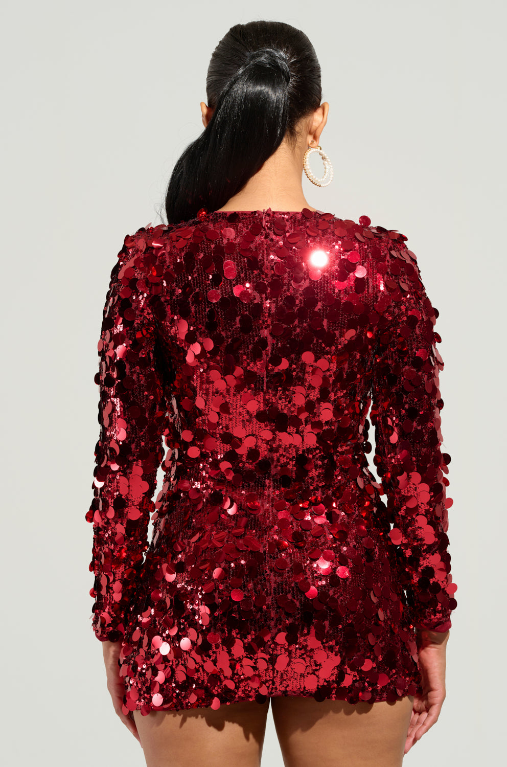 A person with long black hair is facing away from the camera, dressed for a party in the dazzling SHE'S ALL THAT SEQUIN MINI DRESS and large hoop earrings.