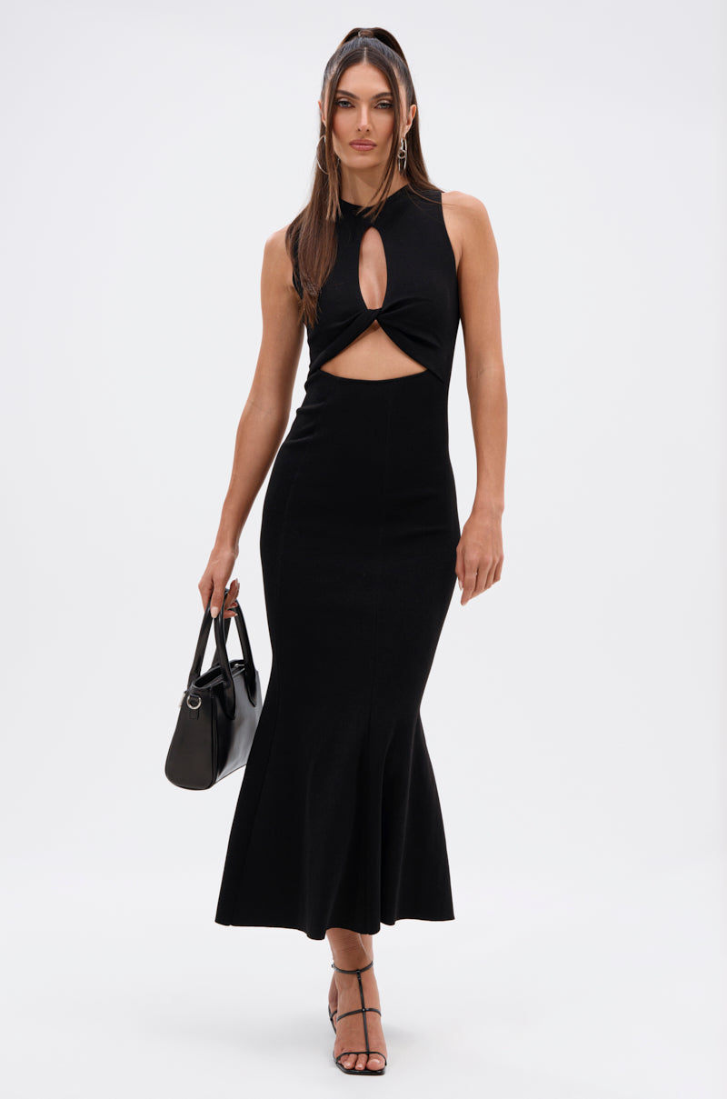 A woman wearing the NOT TOO CASUAL KNIT MAXI DRESS IN BLACK, featuring a sleeveless design, front cutout, and flared hem, stands against a white background with a black handbag and strappy heels.