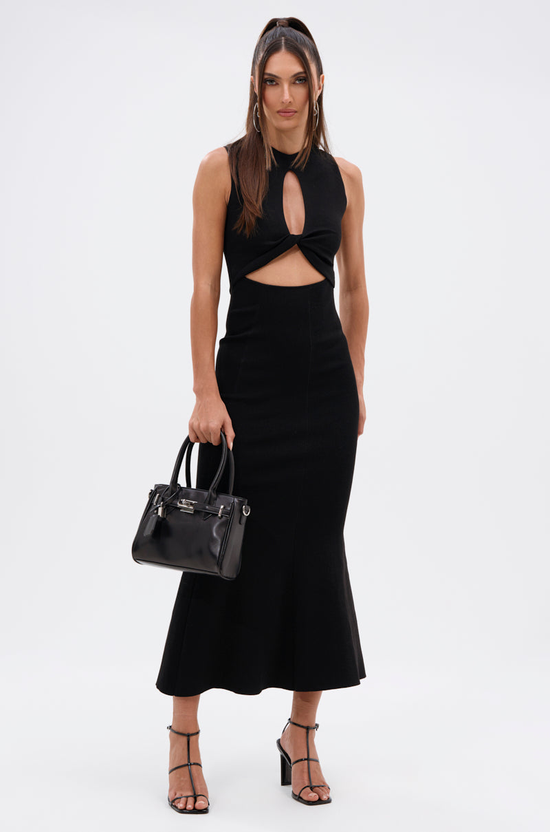 A woman with long, straight hair poses against a white background wearing the NOT TOO CASUAL KNIT MAXI DRESS IN BLACK, paired with a black handbag and strappy black heels.