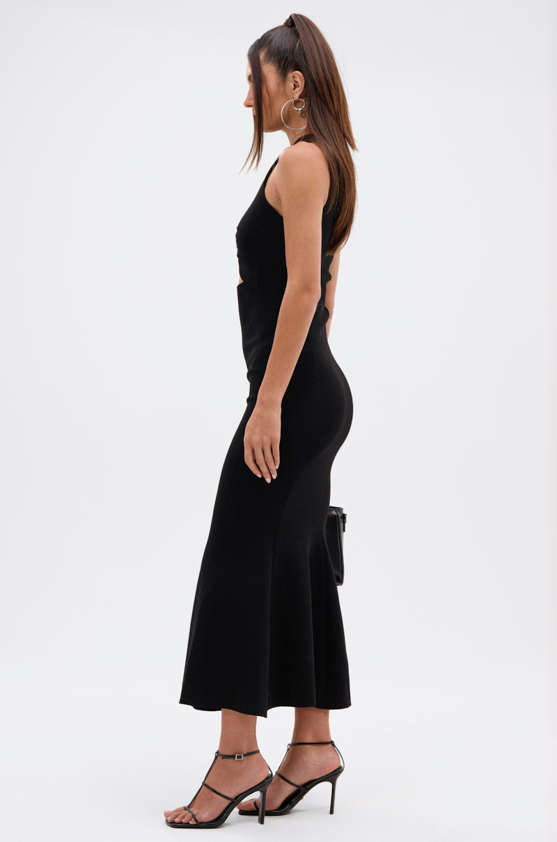 A woman stands in profile wearing the NOT TOO CASUAL KNIT MAXI DRESS IN BLACK, styled with high-heeled sandals, large hoop earrings, a small handbag, and her long brown hair straight against a plain white background.