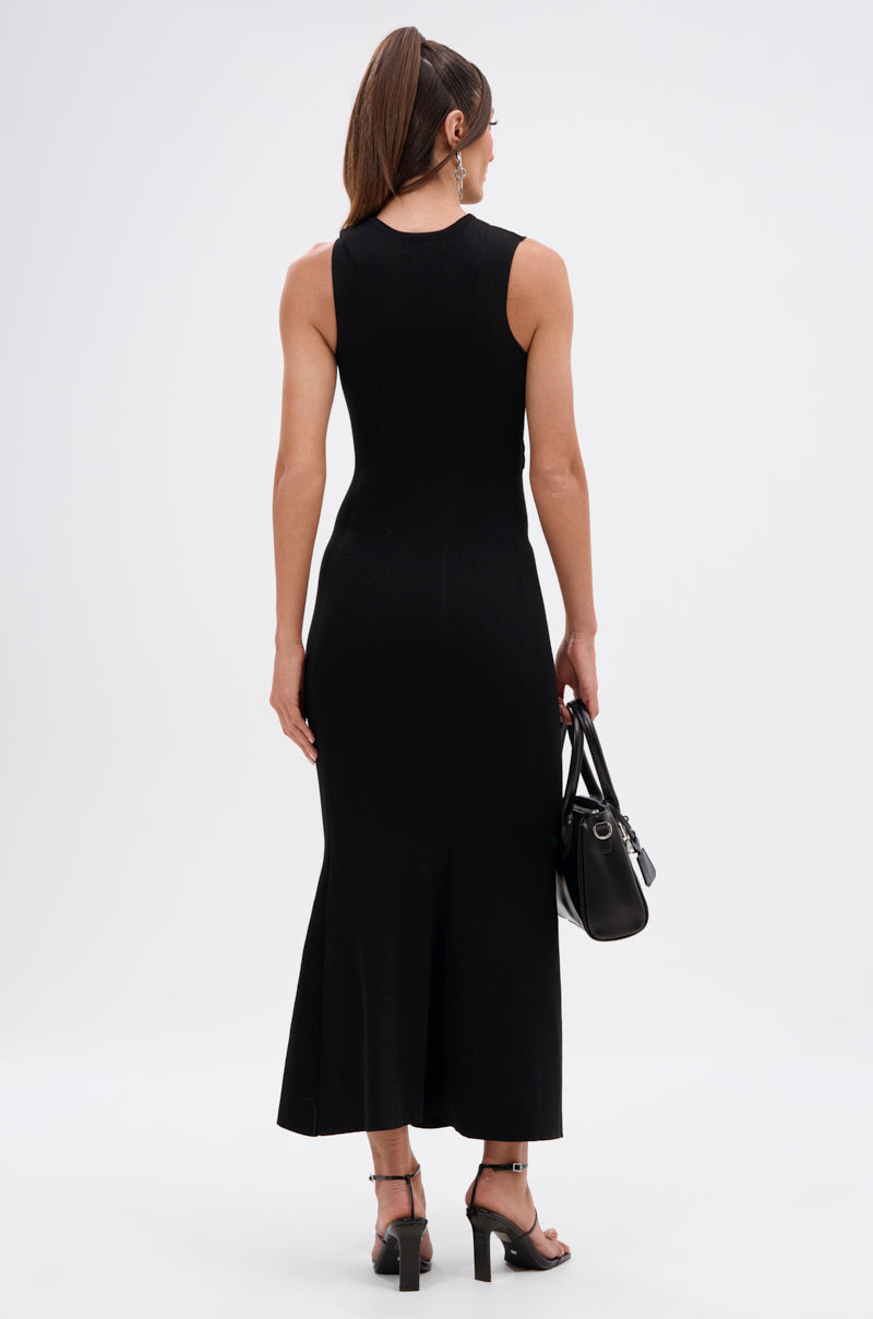 A woman with long brown hair in a ponytail wears the NOT TOO CASUAL KNIT MAXI DRESS IN BLACK, paired with black heeled sandals and a black handbag, standing against a plain white background facing away.