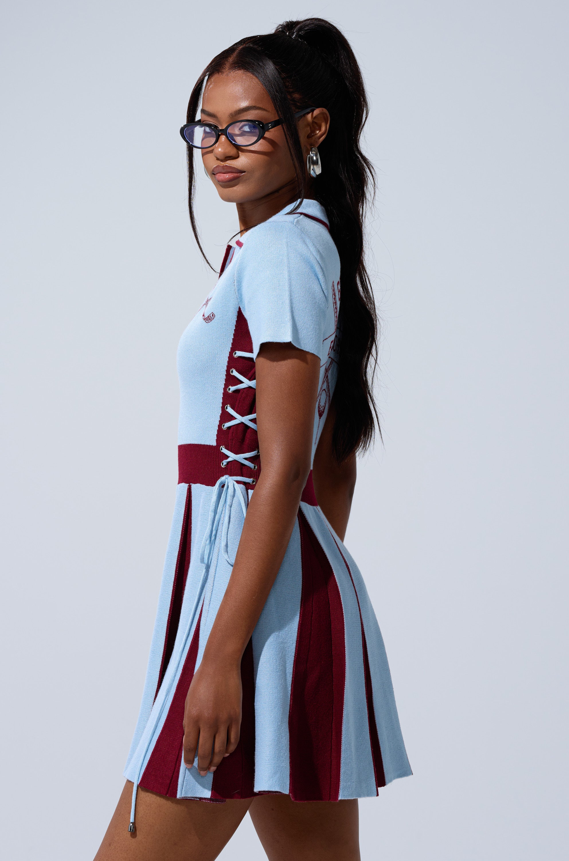 A woman with long dark hair in a ponytail wears glasses and the DREKE KNIT MINI DRESS IN BLUE AND BURGUNDY, featuring short sleeves and contrasting side panels, standing against a plain light background, looking over her shoulder.