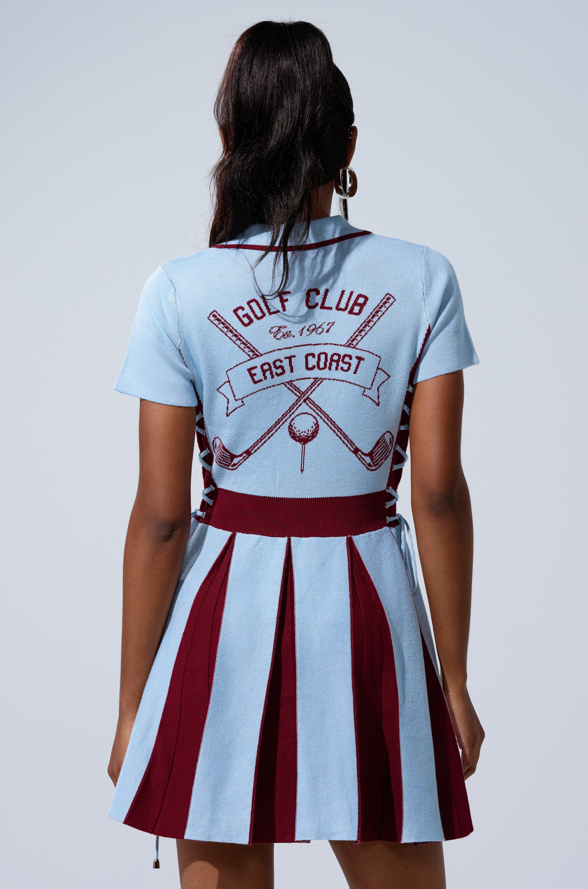 A person with long hair wears the DREKE KNIT MINI DRESS IN BLUE AND BURGUNDY—a light blue and maroon collared polo dress with short sleeves, a preppy pleated skirt, and golf-themed graphics plus "GOLF CLUB Est. 1967 EAST COAST" on the back.