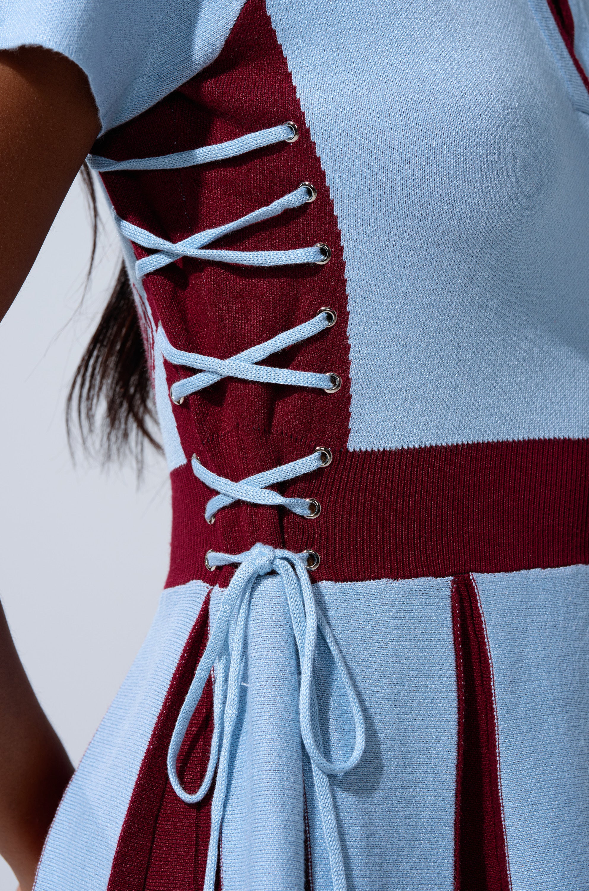 Close-up of a person wearing the DREKE KNIT MINI DRESS IN BLUE AND BURGUNDY, featuring maroon side panels and light blue lace-up detailing with silver eyelets.