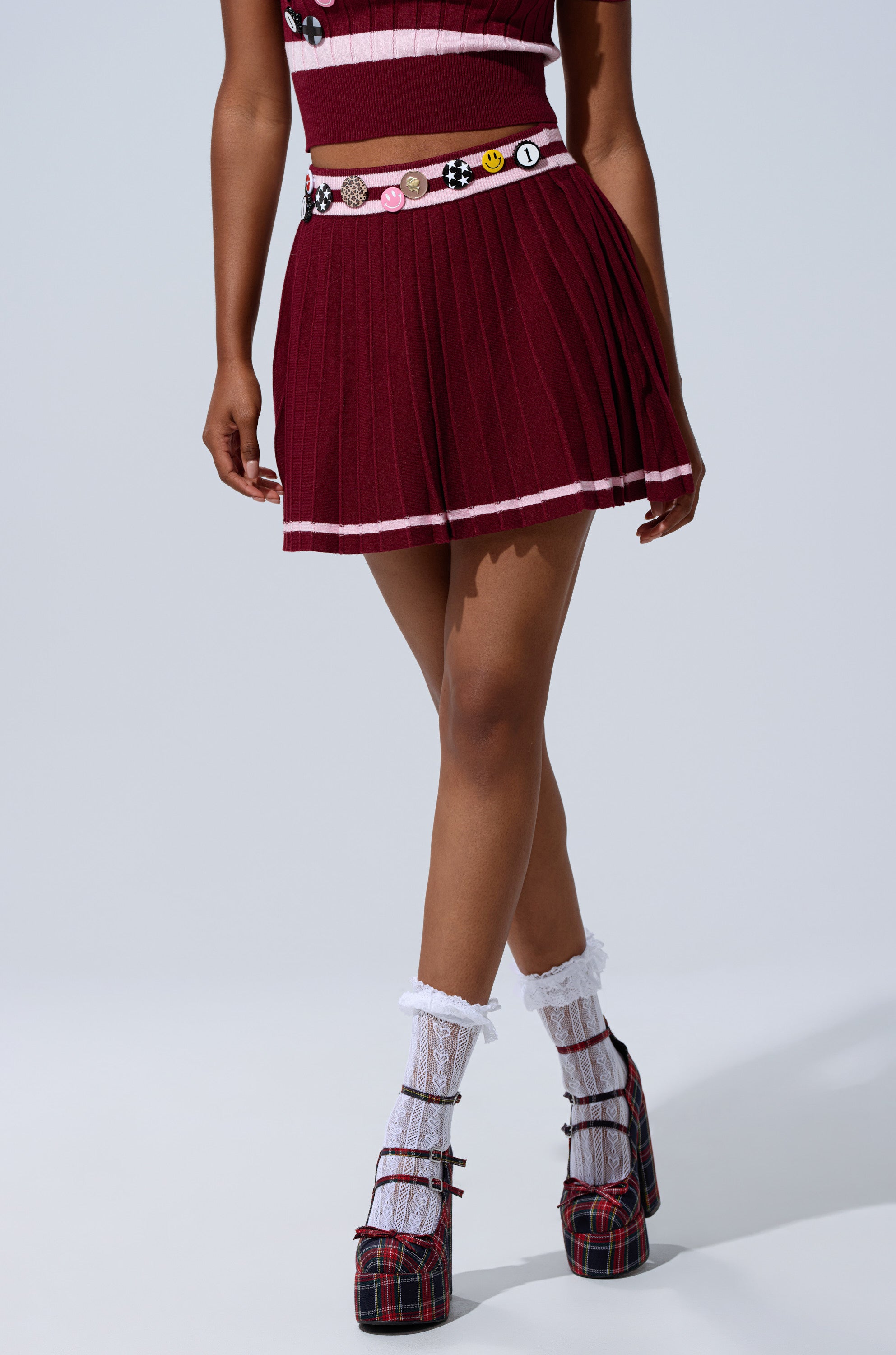 Wearing the DREKE KNIT PLEATED MINI SKIRT in burgundy with a matching crop top, colorful pin belt, white lace socks, and plaid platform heels, a person poses against a plain light background.