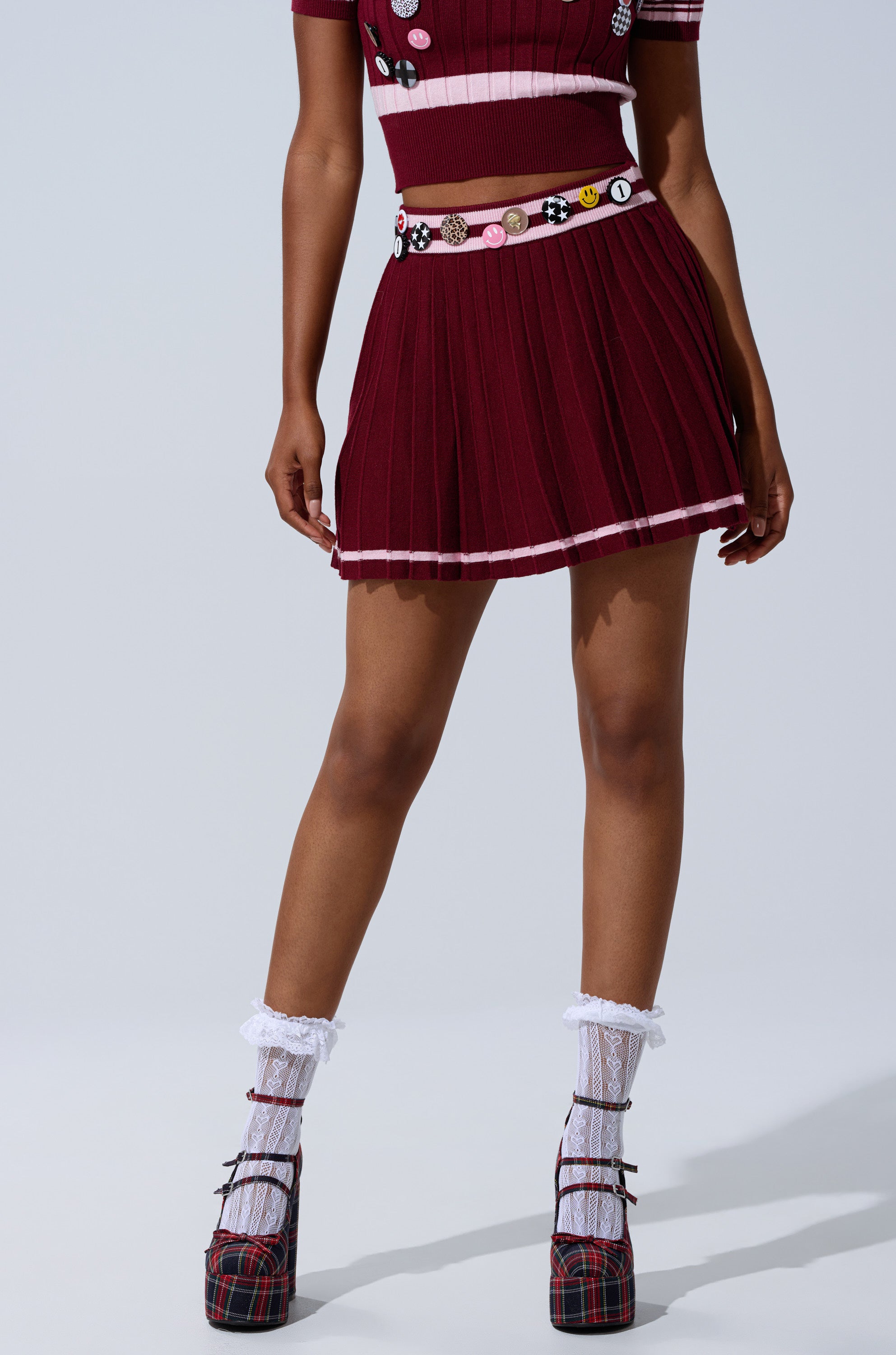 A person wearing a maroon DREKE KNIT PLEATED MINI SKIRT, a matching crop top, white frilly socks, plaid platform heels, and a button-adorned belt stands against a plain light background.