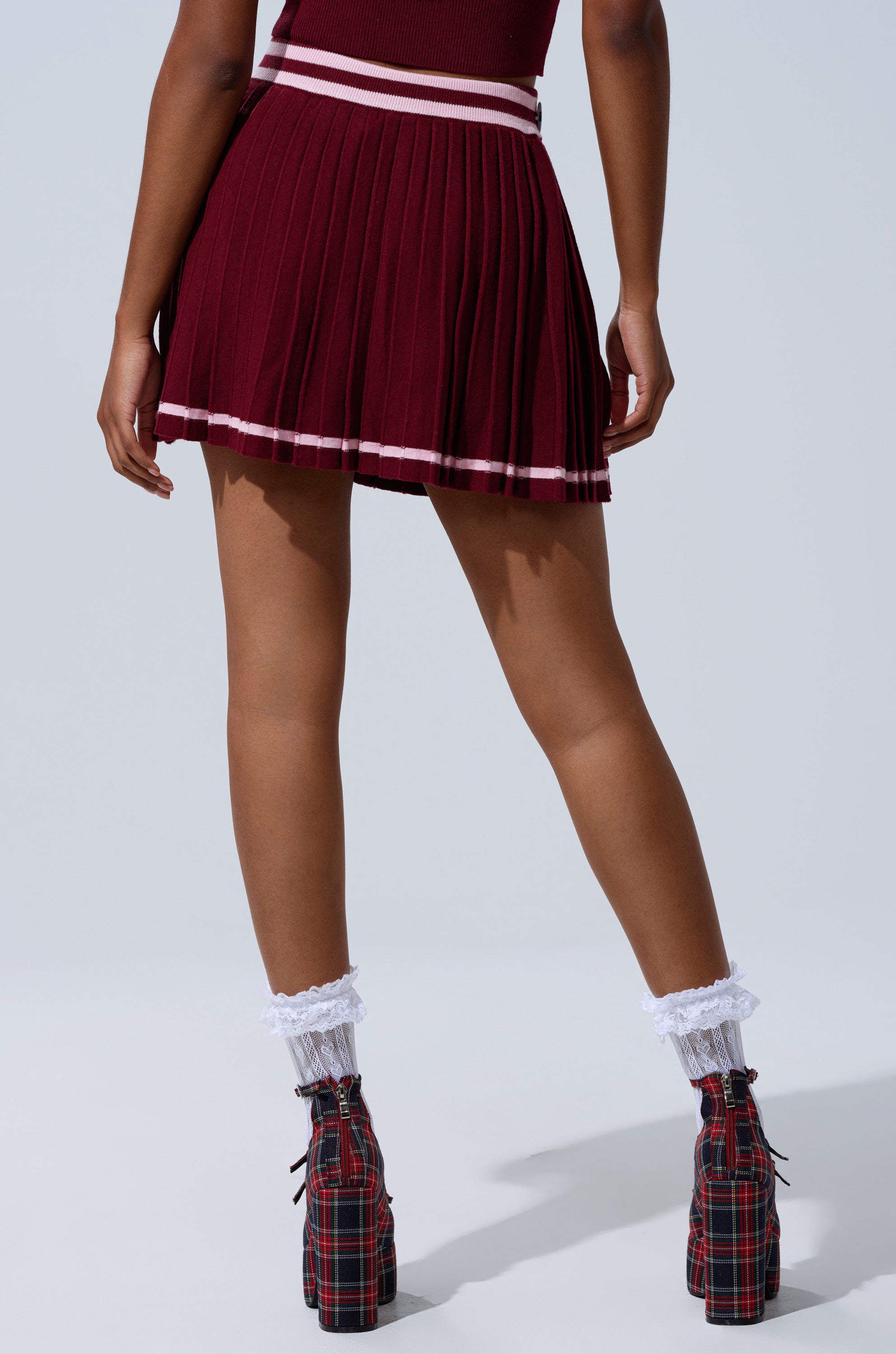 A person stands facing away, wearing the DREKE KNIT PLEATED MINI SKIRT with plaid platform shoes and white lace-trimmed socks, set against a plain light background.