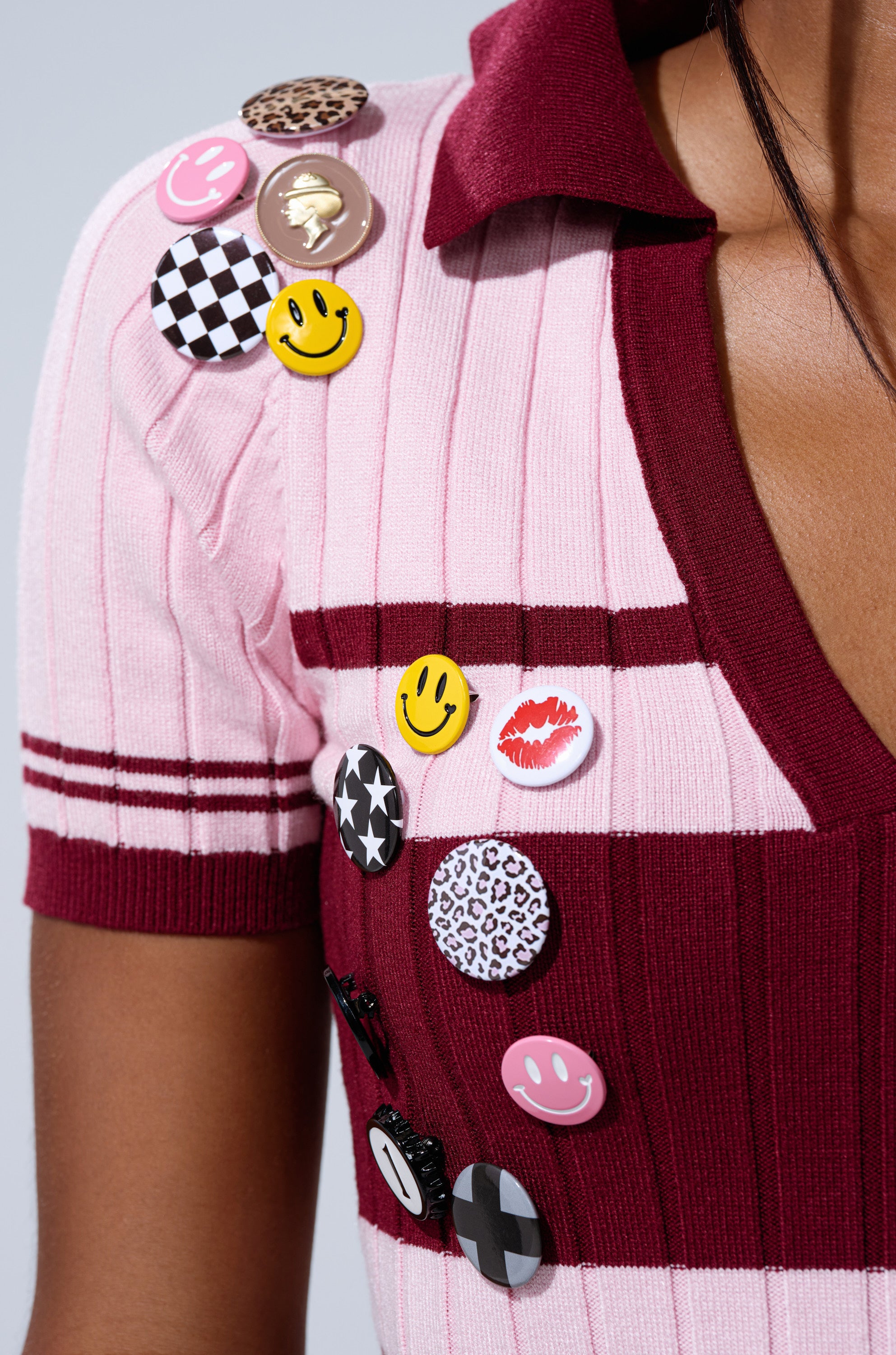 A person wearing the DREKE KNIT POLO, a pink and burgundy ribbed cropped polo decorated with colorful pins—smiley faces, animal prints, and checkered patterns—attached to the shoulder and chest.