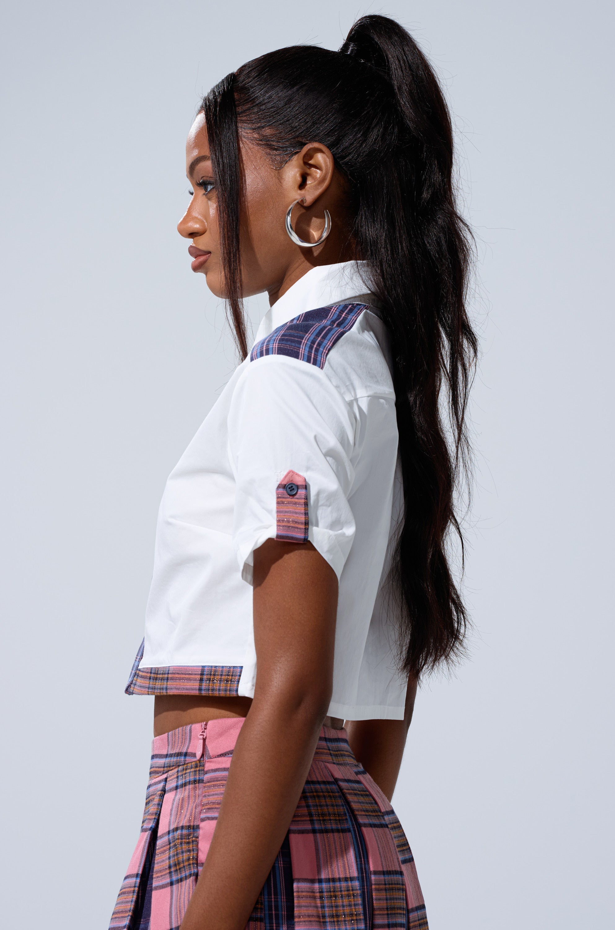 A woman in profile wears the DREKE CROPPED POPLIN SHIRT with plaid accents, paired with matching pink plaid pants and large hoop earrings. Her long, wavy hair is styled in a high ponytail against a plain light gray background.
