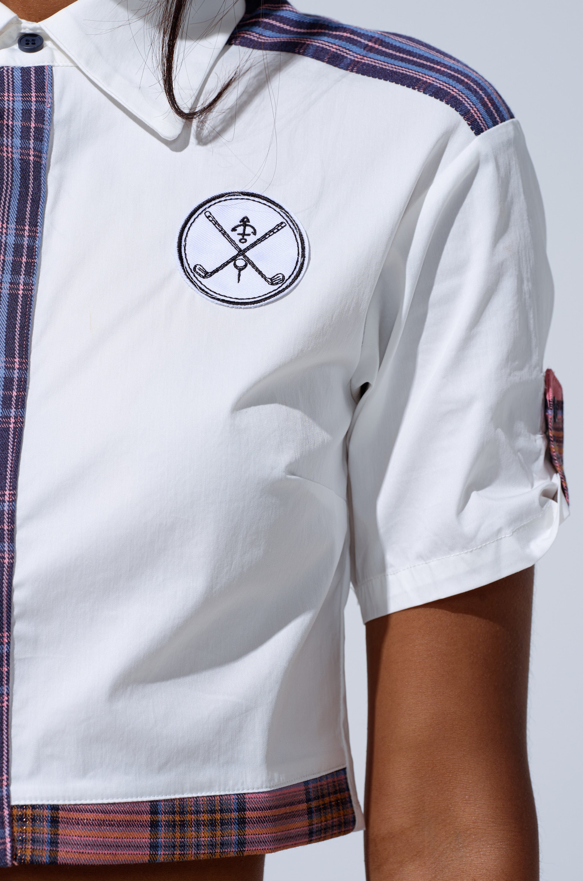A close-up of a person wearing the DREKE CROPPED POPLIN SHIRT, featuring plaid accents and a black embroidered chest patch with crossed objects and a snake for a preppy-chic, stylish edge.