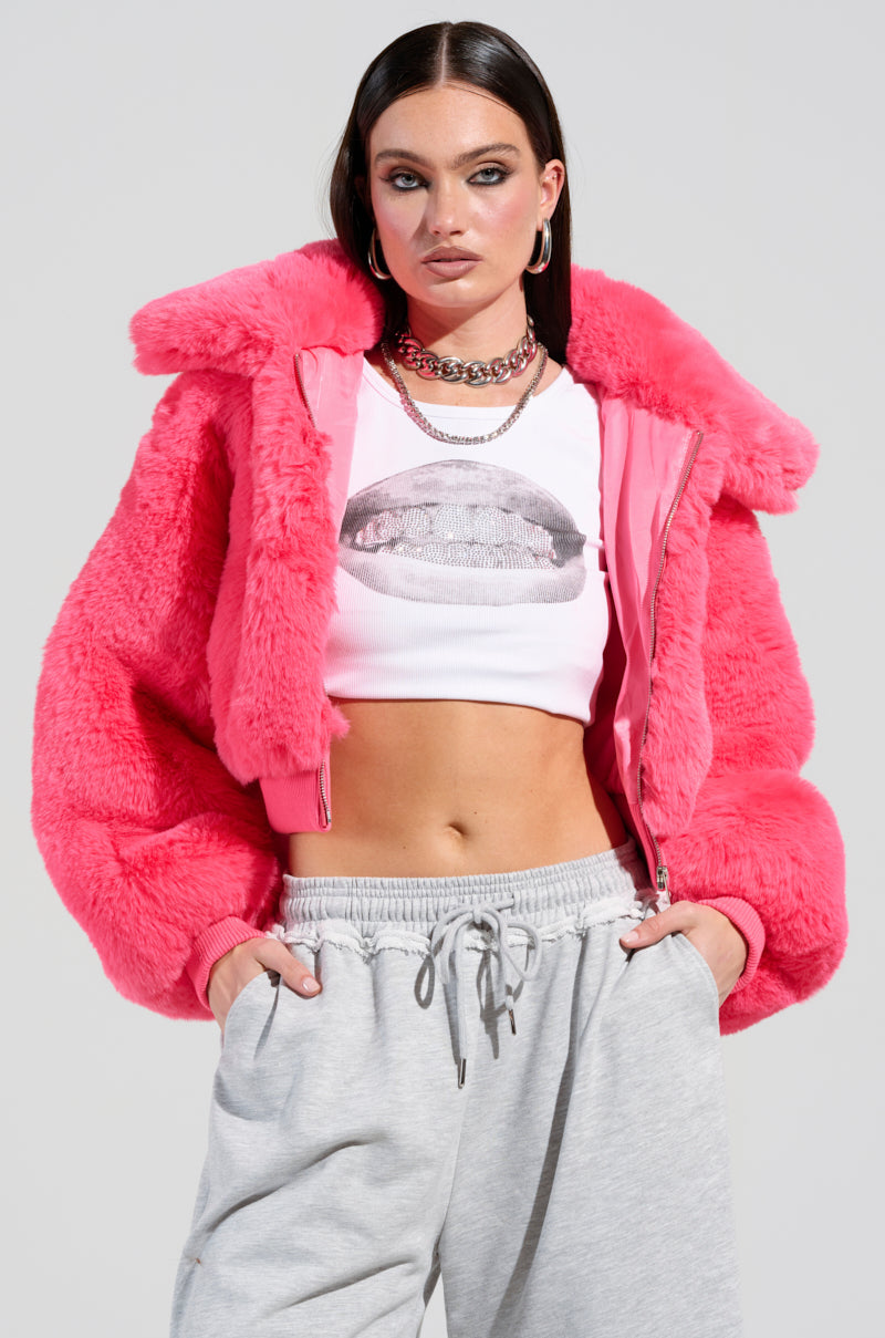 A person confidently poses in the DREW Faux Fur Bomber in Hot Pink layered over a cropped white top featuring a graphic design, paired with gray sweatpants. The look is accessorized with hoop earrings and layered necklaces, all set against a neutral gray background.