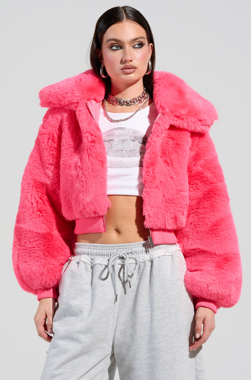 Dressed in the DREW FAUX FUR BOMBER IN HOT PINK over a white crop top, paired seamlessly with gray sweatpants, an individual exudes style. Their long dark hair cascades freely as they accessorize with hoop earrings and a chunky chain necklace, standing confidently against a plain background.