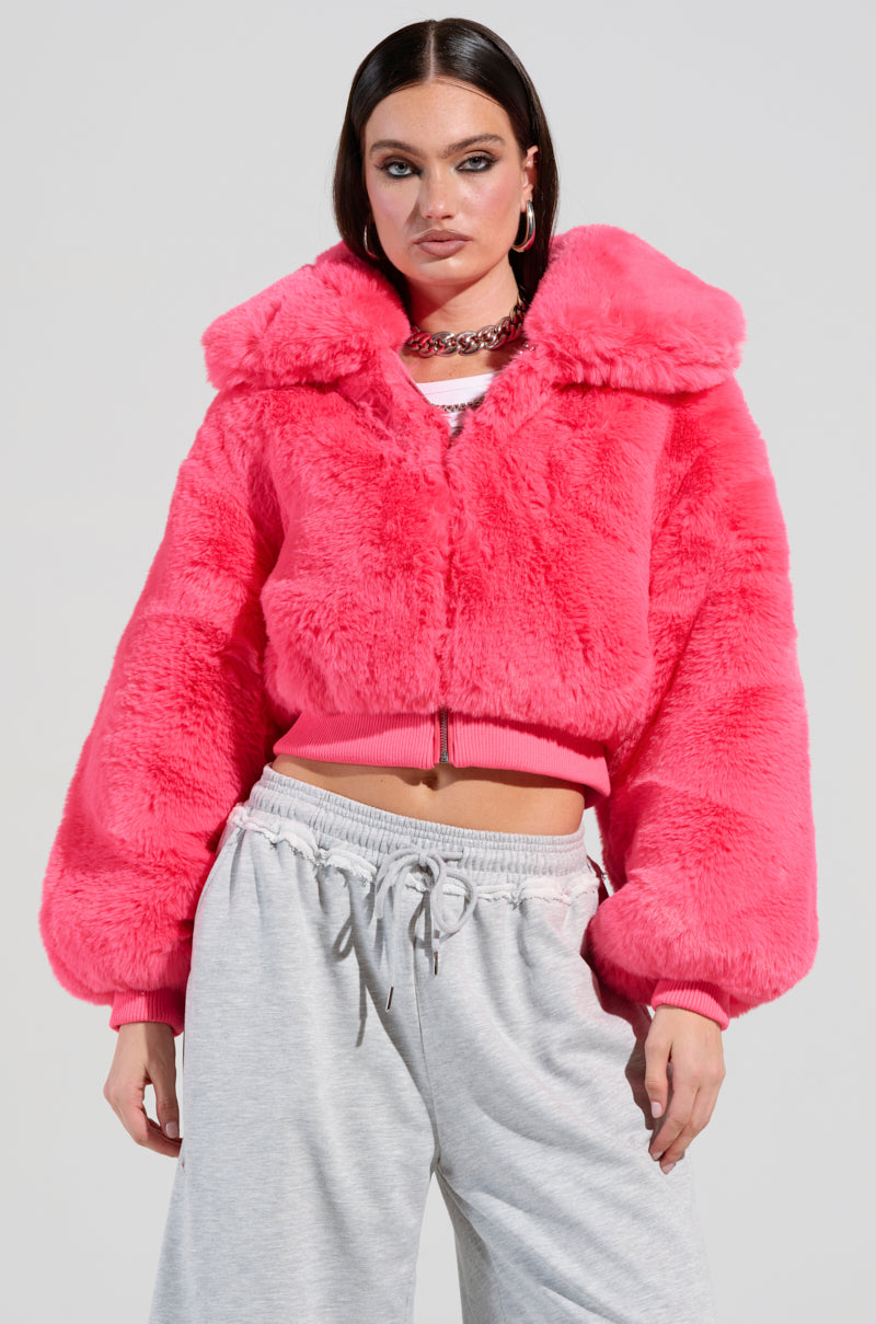 A woman is posing in the DREW Faux Fur Bomber in hot pink, paired with gray sweatpants. Her long dark hair complements her look, enhanced by hoop earrings and a chain necklace, all against a simple background.