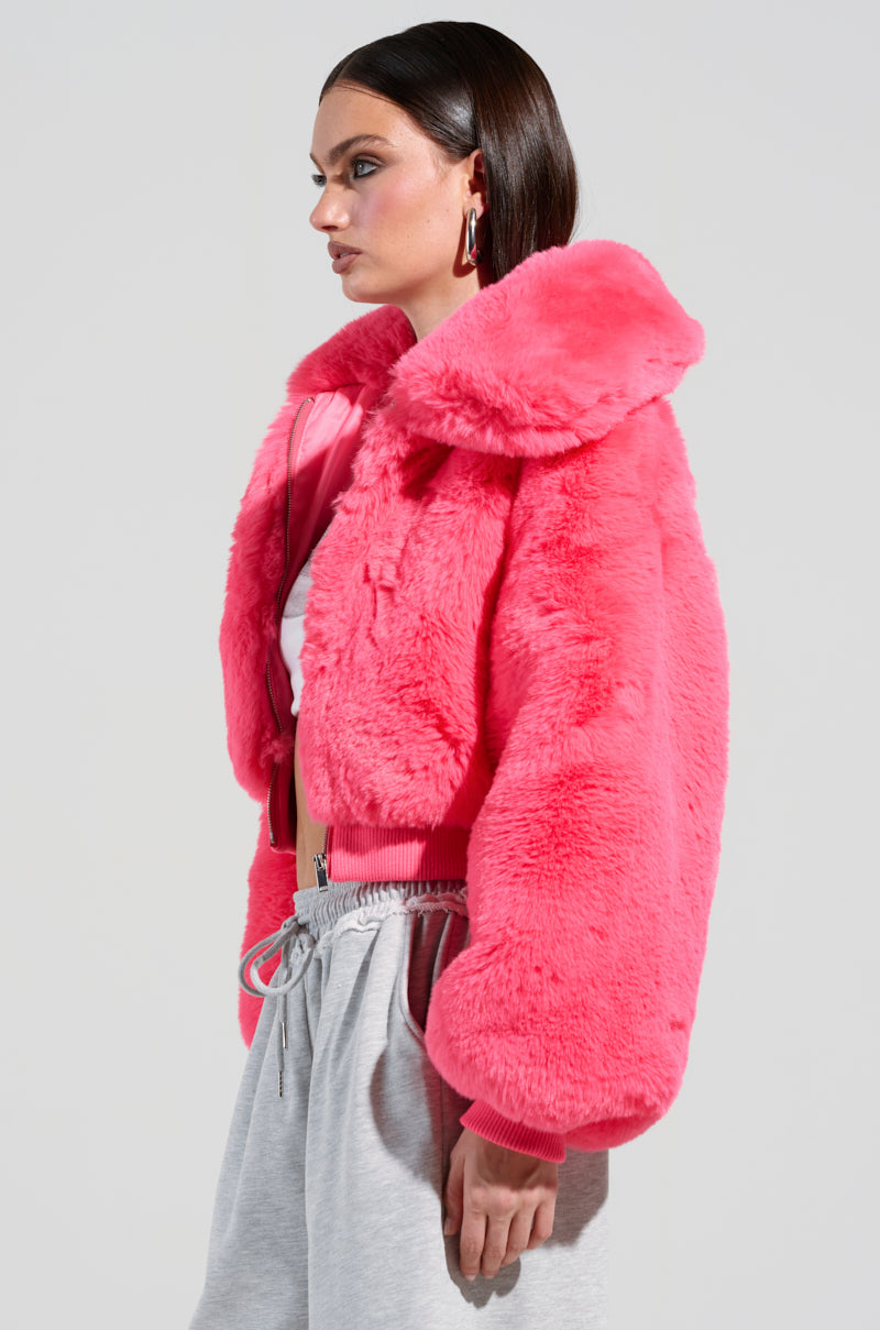 A person dressed in the DREW FAUX FUR BOMBER IN HOT PINK stands against a plain background. The jacket's cropped silhouette features a large collar. They complement the look with a white top and gray sweatpants, completing their stylish ensemble with shoulder-length dark hair.