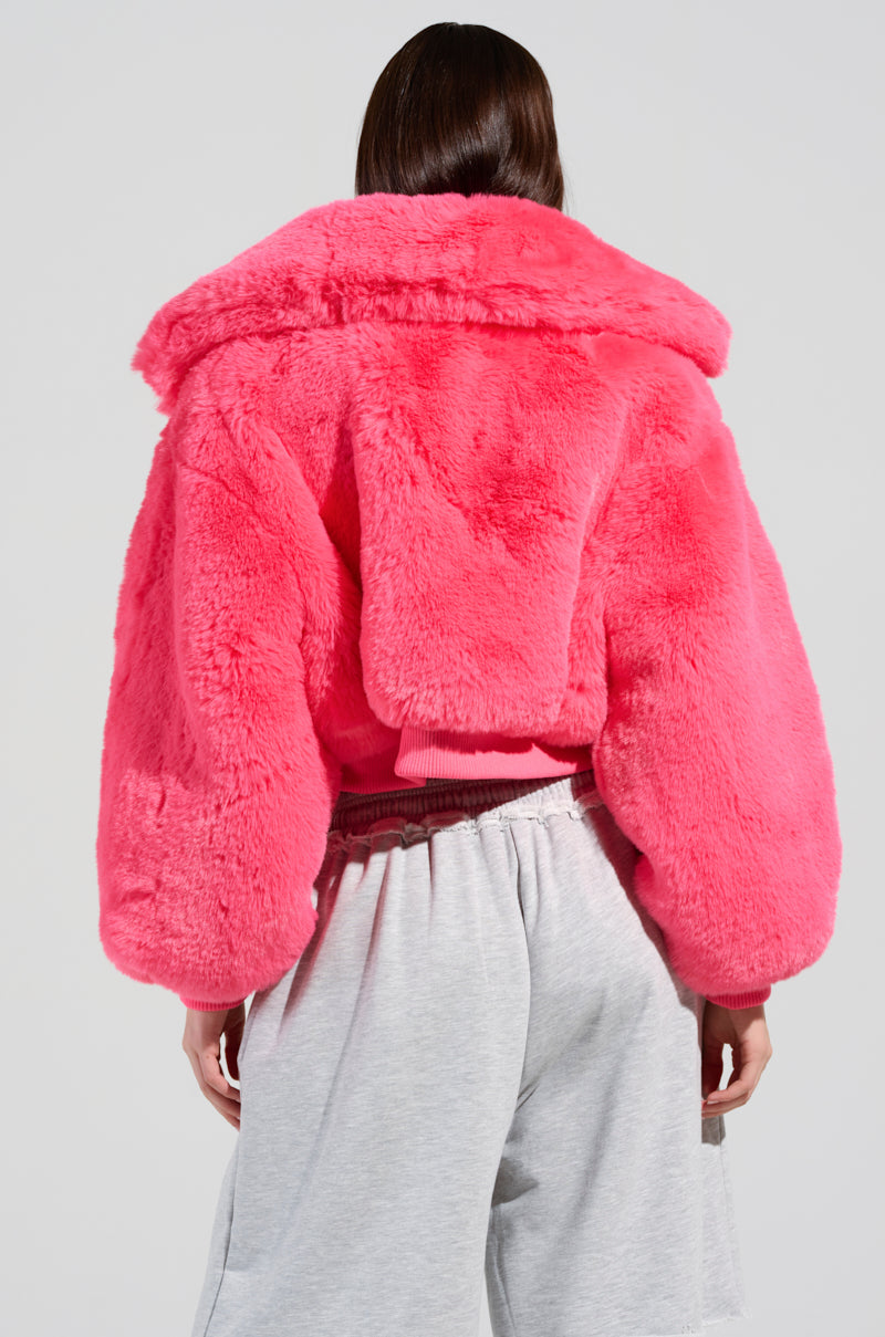 Sporting the Drew Faux Fur Bomber in Hot Pink, a person with their back to the camera showcases a striking look with its large collar and oversized sleeves. Paired with light gray sweatpants, this eye-catching cropped silhouette stands out against a plain background.