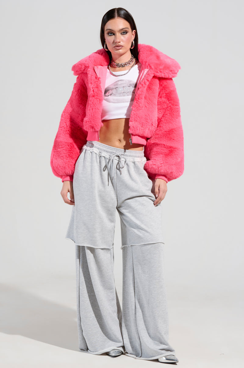 Wearing the DREW FAUX FUR BOMBER IN HOT PINK over a white cropped top and loose gray sweatpants, a person with long dark hair showcases a chic cropped silhouette accentuated by a silver chain necklace. The setting features a plain, light-colored background.