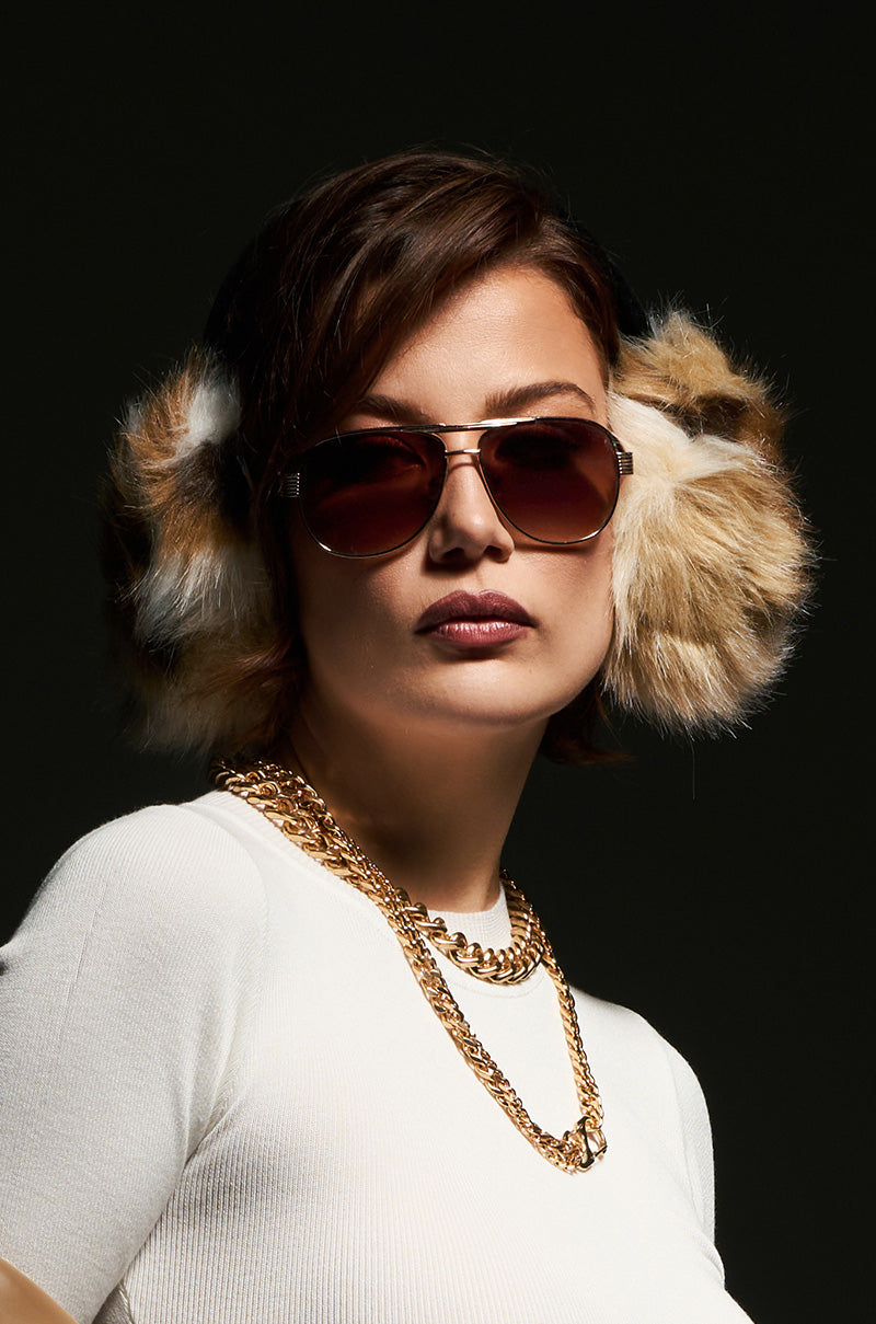 A person with short brown hair is seen wearing large WILD ONE FAUX FUR EAR MUFFS, aviator sunglasses, and a white sweater, complemented by a gold chain necklace. The dark background highlights the fashion-forward winter accessory.