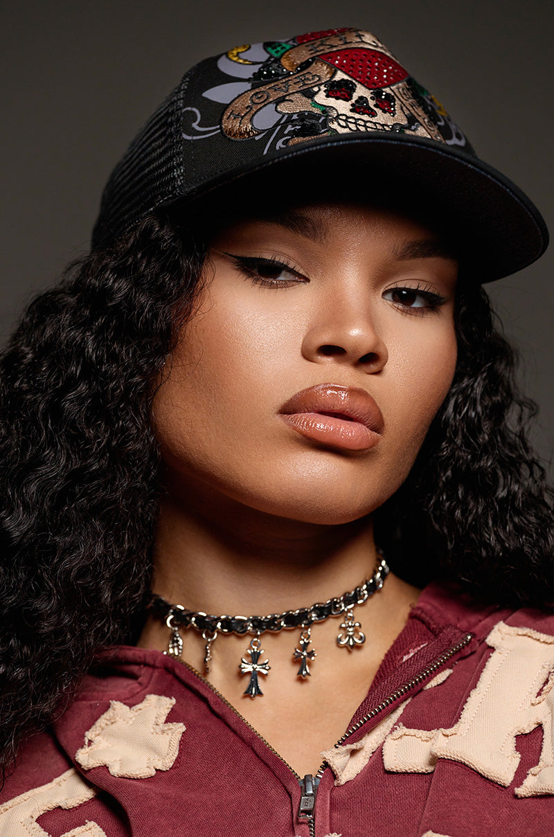 Sporting an ED HARDY RHINESTONE LOVE KILLS SLOWLY HAT embellished with the iconic rhinestone design, a maroon jacket, and a beaded necklace with small pendant charms, the individual with long, curly hair stares directly into the camera against a dark backdrop.