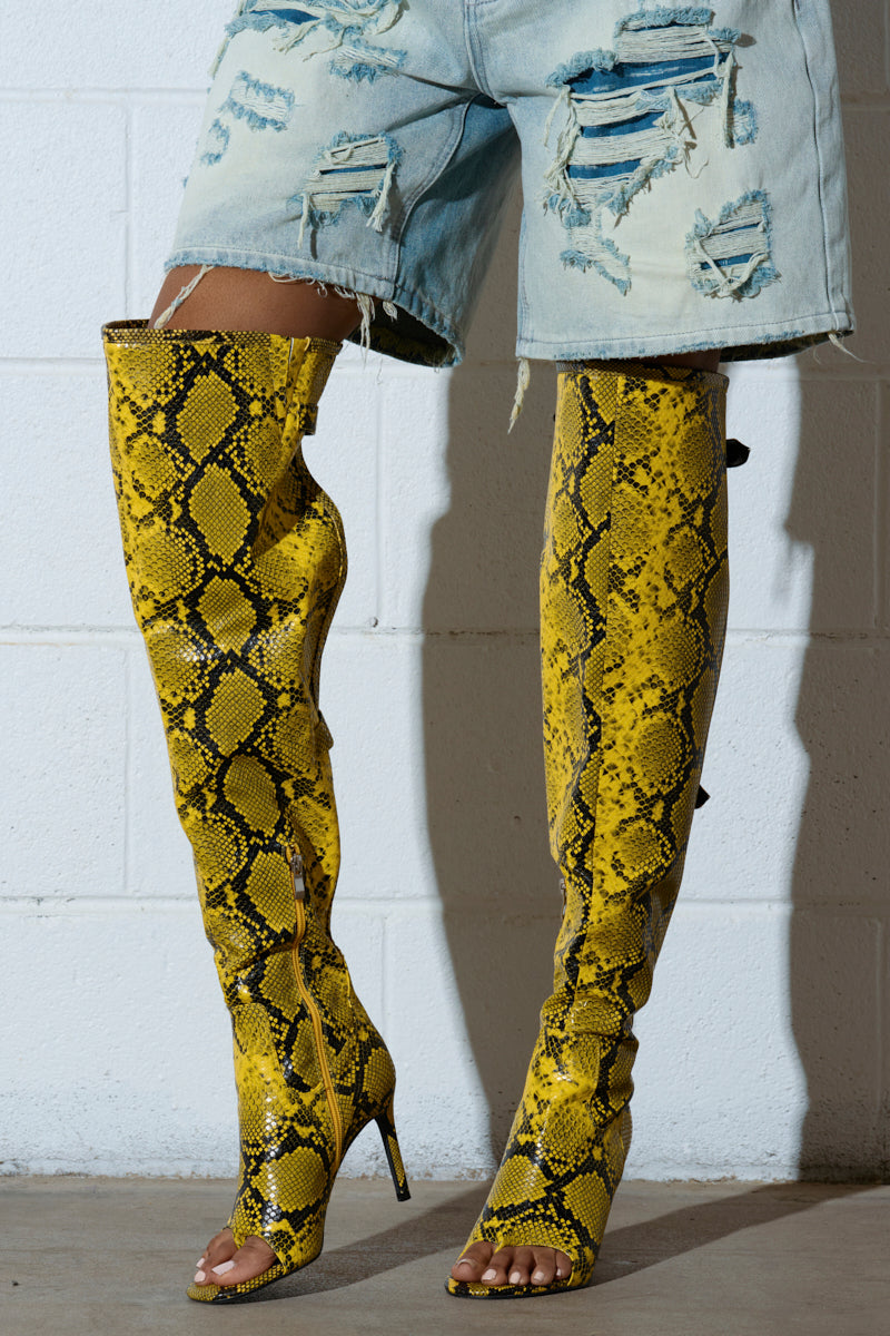 Someone wearing distressed denim shorts and the AZALEA WANG ELIAM BOOT IN YELLOW, featuring thigh-high faux snakeskin leather, open toes, and stiletto heels, stands against a white brick wall.