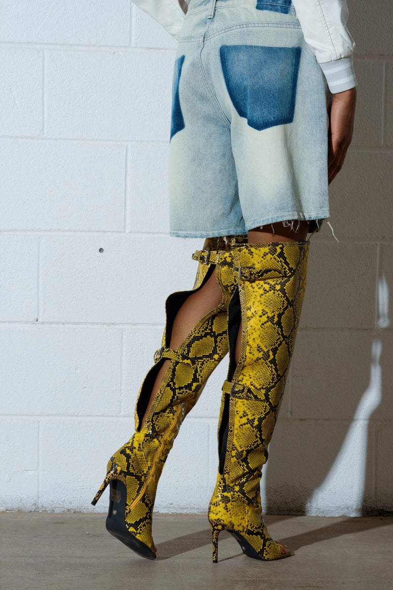 Against a white brick wall, someone wears denim shorts and the AZALEA WANG ELIAM BOOT IN YELLOW, knee-high stiletto boots with faux snakeskin leather texture, black accents, and open back detailing.