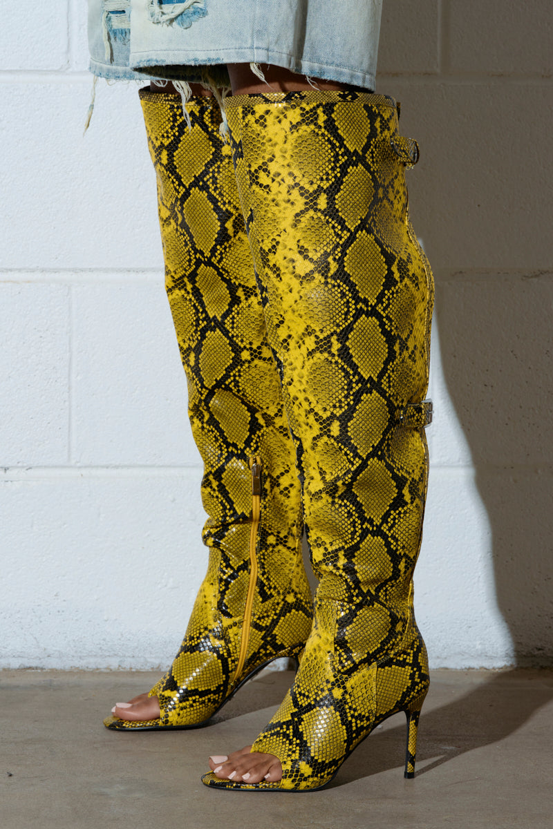 The AZALEA WANG ELIAM BOOT IN YELLOW is showcased on a concrete floor against a white wall. It's a pair of thigh-high faux snakeskin leather stiletto boots with peep toes and side zippers, alongside a skirt featuring a frayed hem.