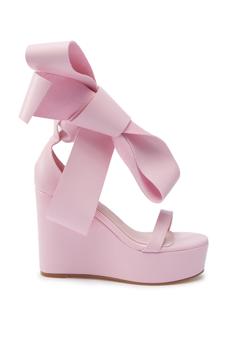 The AZALEA WANG EMEKA Pink Platform Sandal with Bow is a light pink faux leather wedge heel, featuring a large wide pink bow wrapping around the ankle and an open toe design.