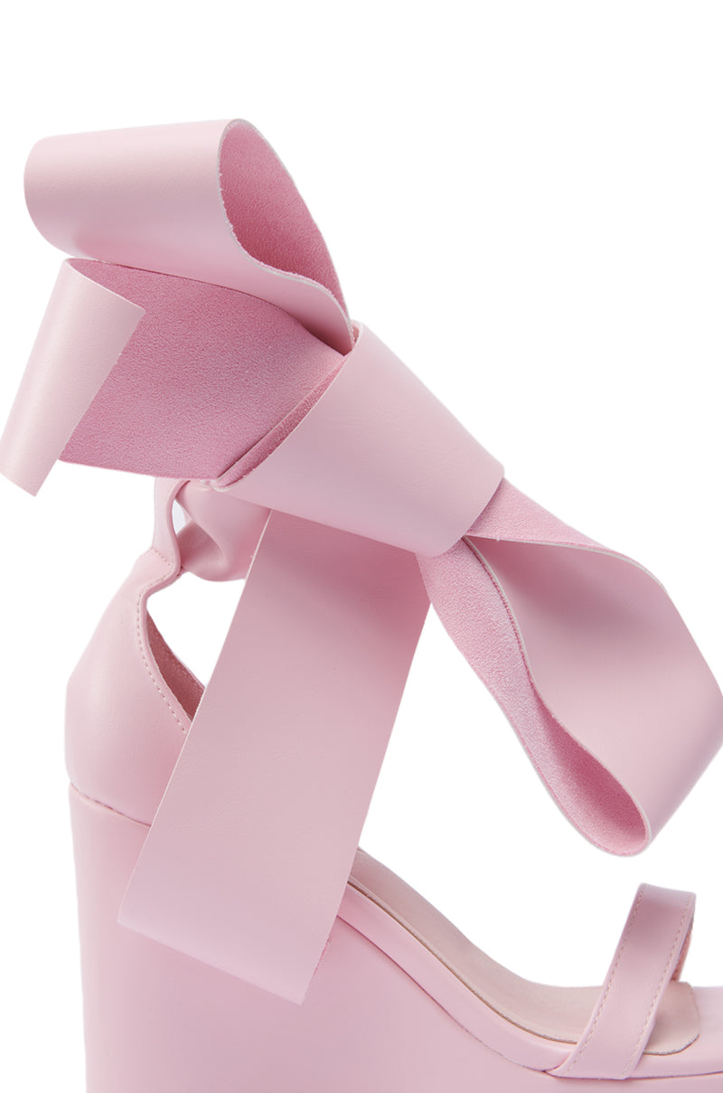 A close-up of the AZALEA WANG EMEKA Pink Platform Sandal, which features a wedge heel and a large decorative pink bow on the faux leather ankle strap.
