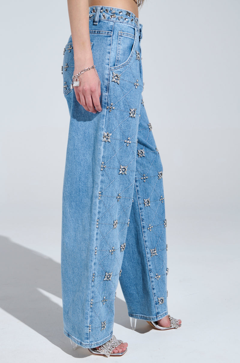 Wearing TAKE ME OUT RHINESTONE DENIM JEANS in light blue with sparkling silver rhinestones, styled with a silver bracelet and open-toe silver heeled sandals against a plain light gray background.
