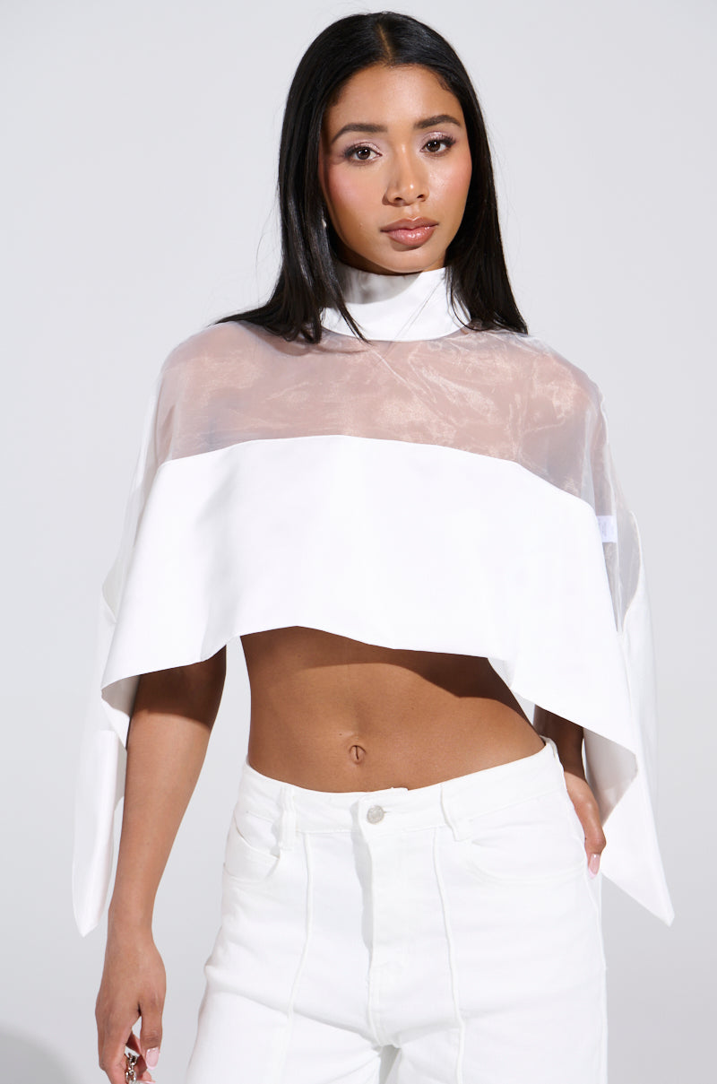 A woman stands confidently against a plain background, wearing the TALK TO ME LATER BLOUSE featuring a sheer mesh bodice with structured shoulders, over a white bandeau and high-waisted white pants. Her long, dark hair is styled straight.