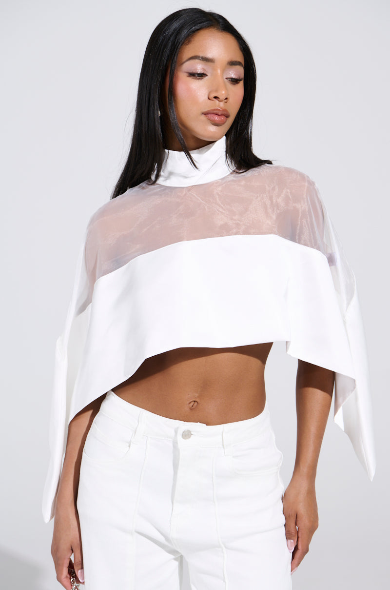 A woman poses gracefully against a plain backdrop, effortlessly stylish. She's wearing the TALK TO ME LATER BLOUSE over a high-neck layer and pairs it with high-waisted white pants. Her long hair cascades freely, while her neutral expression enhances the chic cape-like silhouette of her outfit.